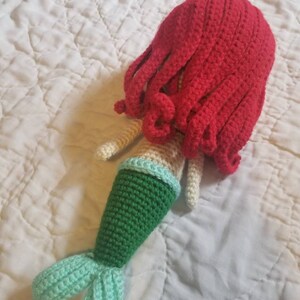 May include: A crocheted mermaid doll with a red hair, green tail, and light blue tail fin.