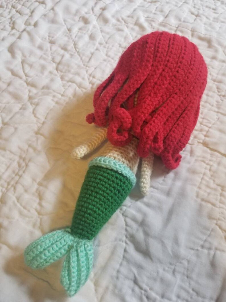 May include: A crocheted mermaid doll with a red hair, green tail, and light blue tail fin.