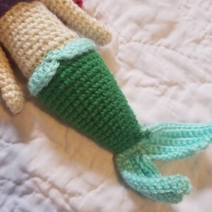 May include: Crocheted mermaid doll with a green tail, a light blue tail fin, and a cream-colored bodice with a purple top.