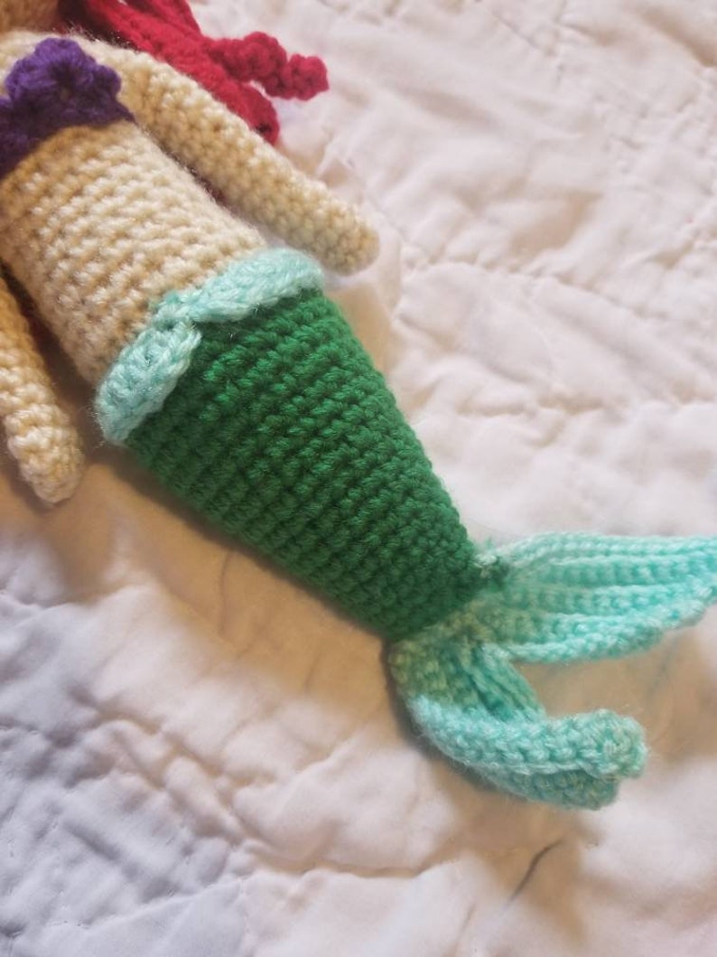 May include: Crocheted mermaid doll with a green tail, a light blue tail fin, and a cream-colored bodice with a purple top.