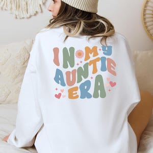 Könnte beinhalten: Weißer Pullover mit dem Text "In My Auntie Era" in bunten, Retro-Lettern. Der Text ist von kleinen Herzen und Sternen umgeben.
