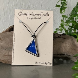 Puede incluir: Un colgante de triángulo de vidrio azul en un collar de cordón negro. El colgante está engastado en plata y tiene una superficie texturizada. El collar está hecho a mano por Myr. La etiqueta dice "GlassAndWoodCrafts Triangle Pendant" e incluye la dirección del sitio web "www.etsy.com/shop/glassandwoodcrafts".