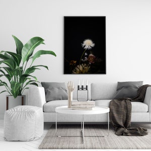 May include: A black and white photo of a single dandelion with other flowers in the background, framed and hanging on a white wall above a gray couch with pillows and a throw blanket.