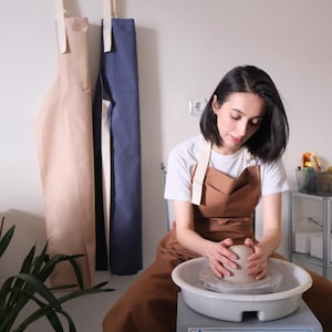 Peut inclure: Une personne façonne de l'argile sur un tour de potier. Elle porte un tablier marron sur un t-shirt blanc. Deux tabliers sont accrochés au mur derrière, un beige et un bleu. Une étagère avec des fournitures de poterie est visible en arrière-plan.