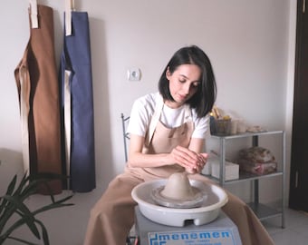 May include: A pottery wheel with a lump of clay being shaped by a person wearing a beige apron. Two aprons, one brown and one blue, hang on the wall. A shelf with items is in the background.