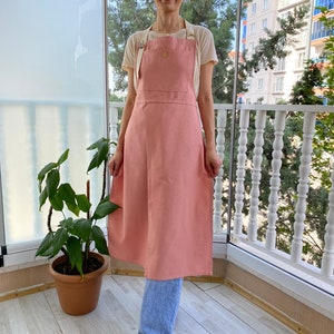 May include: A long pink linen apron with adjustable straps. The apron is worn over a white t-shirt and blue jeans.