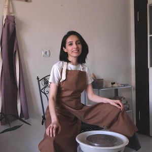 May include: A woman wearing a brown apron with cream straps, sitting near a pottery wheel. A purple apron hangs on the wall. The woman is smiling. The pottery wheel is white.