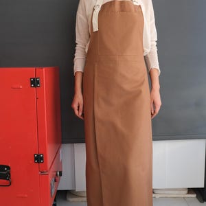 Op de afbeelding: Een lange, bruine schort met een borstzak en witte bandjes. De schort wordt over een lichtgekleurde trui gedragen. Op de achtergrond is een rode oven te zien.