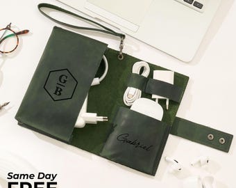May include: Dark green leather tech organizer with a wrist strap, open to reveal white charging cables, a charger, and a wireless mouse. The organizer has a monogram "GB" and the name "Gabriel" on it. A laptop and earbuds are also visible.