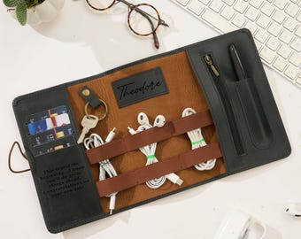 May include: Black and brown leather tech organizer with a personalized nameplate that reads "Theodore." The organizer holds cards, keys, and various tech accessories. Includes a zipper pocket and a pen holder. Text on the organizer reads: "This is just the beginning..."