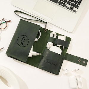 May include: Open green leather tech organizer with a wrist strap, holding white charging cables, a charger, and earbuds. The organizer has a snap closure and a pocket with the name "Gabriel" printed on it. The front has the initials "G B" inside a hexagon.