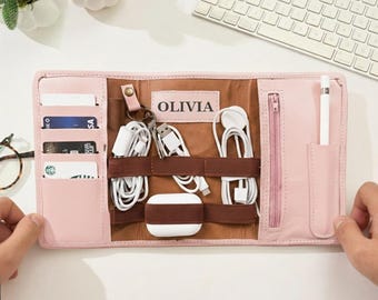 May include: A pink leather organizer with multiple compartments. It features card slots, a zippered pocket, and elastic bands to hold cables and accessories. The name "OLIVIA" is printed on a brown patch. A white stylus and various electronic accessories are visible.