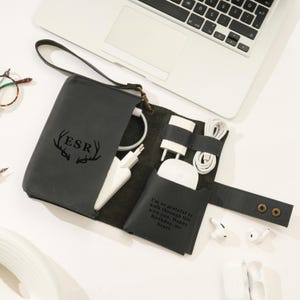 May include: A dark gray leather tech organizer with a wrist strap, open to reveal white charging accessories. The organizer has the initials "ESR" and a deer antler design. A quote is printed on the inside. A laptop and earbuds are also visible.