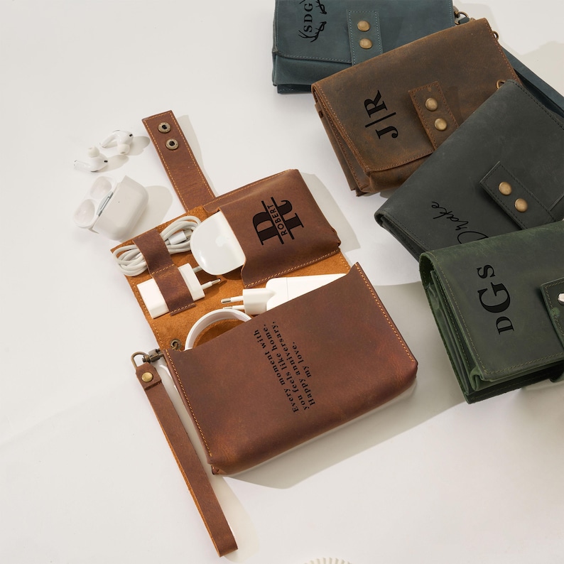 May include: A collection of personalized leather tech organizers in various colors, including brown and green. The organizers are open, displaying charging cables and a charger. Some have initials or names engraved, and one has a quote.