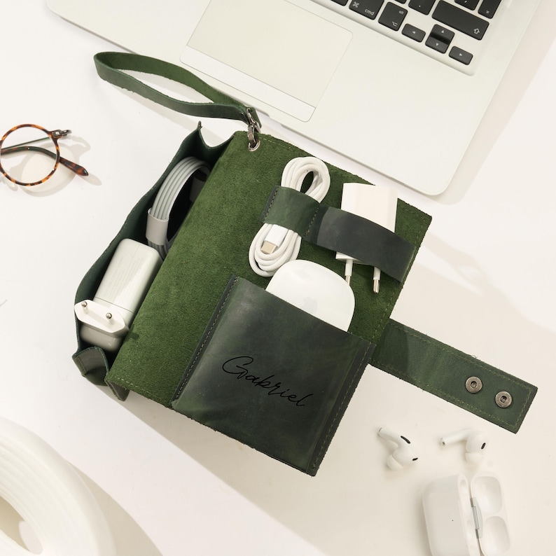 May include: A green leather tech organizer with a handle, holding various white charging cables, a mouse, and a charger. The organizer has a pocket with the name "Gabriel" embossed on it. A laptop, glasses, and wireless earbuds are also visible.