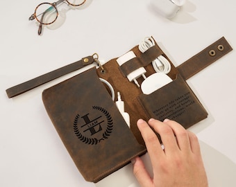 May include: A brown leather tech organizer with a wrist strap and snap closure. The front features a laser-engraved monogram with the name LIAM inside a laurel wreath. The interior contains slots holding a white power adapter and coiled charging cable.