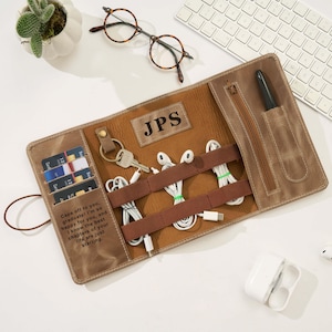 May include: A brown leather roll-up organiser with multiple compartments. It features a zipped pocket, card slots, and straps to hold cables and accessories. The organiser is personalised with the initials "JPS" and includes the text "Caps off to you, graduate!"