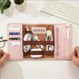 May include: A light pink leather organizer case featuring the name OLIVIA printed in the center. The interior includes card slots, elastic bands for white charging cables, a small zippered pocket, and a dedicated sleeve for a white stylus pen.