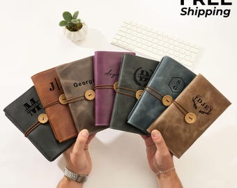 May include: A collection of personalized leather journals in various colors, including gray, brown, and purple. Each journal is secured with a leather cord and wooden button closure and features custom engraved initials or names.