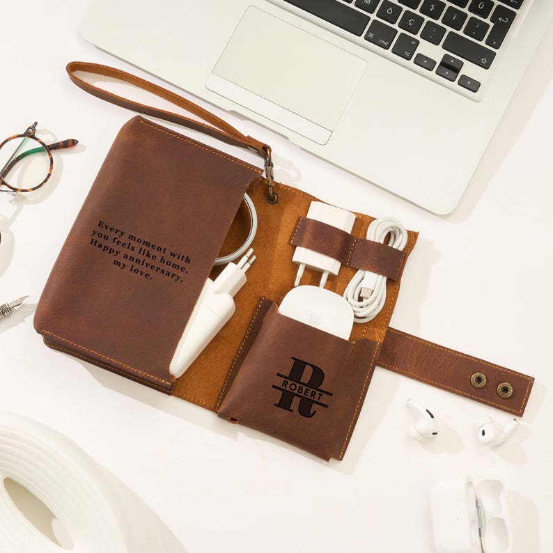 May include: A brown leather tech organizer with a wrist strap, holding a charger, cables, and a small pouch with the name "ROBERT". The organizer has the text "Every moment with you feels like home. Happy anniversary, my love."