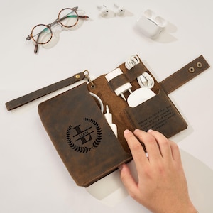 May include: A brown leather tech organizer with a wrist strap, open to reveal compartments for chargers and accessories. The organizer is personalized with the name "LIAM" and a laurel wreath design. Text on the inside reads "Wherever life takes you..."