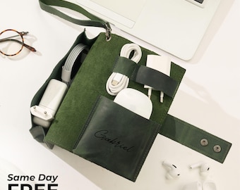 May include: Green leather tech organizer with multiple compartments holding a charger, headphones, and earbuds. The organizer has a strap and snap closure. The name "Gabriel" is embossed on a small pocket. A laptop and glasses are in the background.
