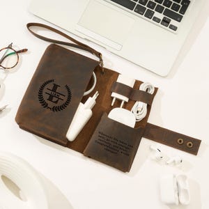 May include: A brown leather tech organizer with a wrist strap, open to reveal chargers and cables. The front is embossed with a monogram and the name "LIAM". A small pocket displays the text: "Wherever life takes you next... Congratulations, world changer!"