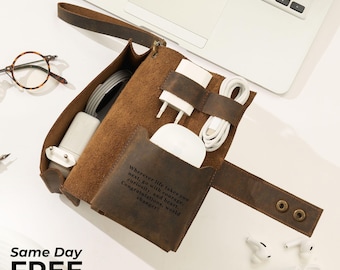 May include: A brown leather tech organizer with a strap, holding white chargers, cables, and a wireless mouse. The organizer has a pocket with the text "Wherever life takes you... world changer!" and is next to a laptop and glasses.