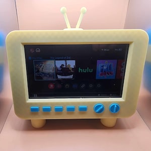 Op de afbeelding: Een gele speelgoedtelevisie met een blauw scherm waarop een videospelmenu wordt weergegeven. Het scherm toont de tekst "The Legend of Zelda" en pictogrammen voor "Zelda", "Demon Survive", "Hulu" en "Fall Guys". De televisie heeft blauwe knoppen en een blauwe knop aan de voorkant.