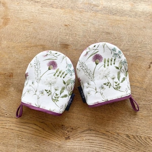 Pode incluir: Um par de luvas de forno brancas com um padrão floral em tons de verde, roxo e branco. As luvas têm uma borda bordô e um laço de suspensão bordô.