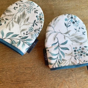 Può includere: Due guanti da forno blu turchese con sfondo bianco e un motivo floreale in tonalità di verde e grigio. I guanti hanno un bordo blu turchese e un anello per appenderli.