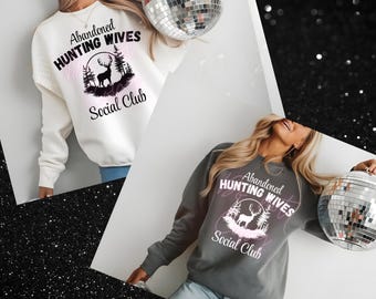 Op de afbeelding: Twee sweatshirts, één wit en één grijs, elk met de tekst "Abandoned Hunting Wives Social Club" en een hertenafbeelding. Het witte sweatshirt wordt gecombineerd met een blauwe jeans. Een discobal wordt op de achtergrond vastgehouden.