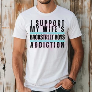 May include: White crew neck t-shirt with the slogan "I SUPPORT MY WIFE'S BACKSTREET BOYS ADDICTION". The text is in black, pink, and turquoise. The shirt is made of cotton and the text is centered on the front.