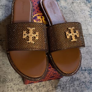 May include: A pair of brown slide sandals featuring a wide strap covered in small bronze-colored crystals and a gold-toned Tory Burch logo emblem. The footbed is brown with the Tory Burch brand name embossed on the heel area of each sandal.