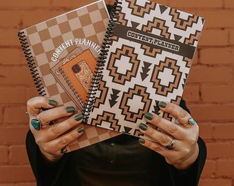 May include: Two spiral-bound "CONTENT PLANNER" notebooks. One has a brown and white geometric pattern, the other a checkerboard design. The notebooks are held in front of a brick wall. The person holding the notebooks is wearing rings.