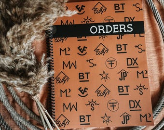 May include: A spiral-bound notebook with a brown cover featuring various black graphic designs and the word "ORDERS" in white. The notebook is surrounded by decorative elements, including dried plants and rope.