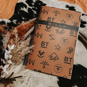 May include: A spiral-bound notebook with a brown cover featuring various black brand symbols and the word "BILLS" in the center. The notebook is open, revealing the patterned design. The background includes dried plants and a cowhide rug.