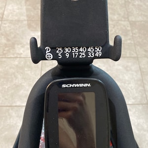 May include: Black exercise bike with a digital display screen and a resistance knob with numbers 25, 30, 35, 40, 45, 50, 5, 9, 17, 25, 33, 49. The screen has a Bluetooth symbol and an up arrow.