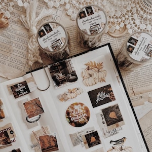 Può includere: Una pagina di scrapbook aperta con adesivi e rotoli di nastro adesivo washi a tema autunnale. Gli adesivi presentano zucche, fogliame autunnale e testo come "Hello Autumn". I rotoli di nastro washi sono etichettati "albumlovehandmade".