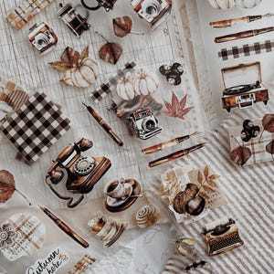 Può includere: Una collezione di adesivi decorativi con illustrazioni a tema autunnale tra cui zucche, fotocamere vintage, penne stilografiche, un telefono a disco, una macchina da scrivere, tazze da caffè e motivi a quadri, con il testo Autumn is here.