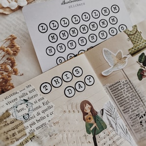 Puede incluir: Una hoja de papel blanco con pegatinas circulares negras que presentan las letras L, M, N y O. Las letras están en una fuente de máquina de escribir. La hoja de papel está encima de un cuaderno con una página que dice "THIS DAY" en pegatinas circulares negras.