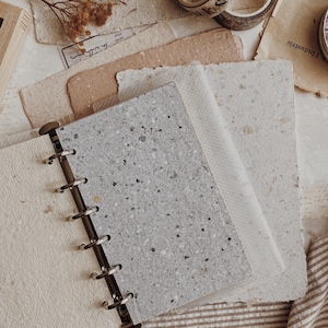 May include: A disc-bound notebook featuring a speckled gray cover with a textured finish, held together by a black metal spine. The notebook is surrounded by various sheets of handmade paper, rolls of decorative tape, and dried floral stems.