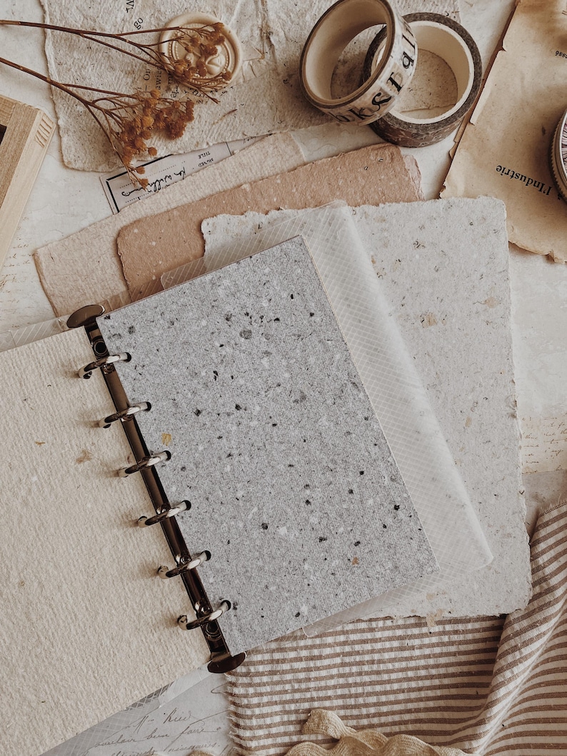 May include: A disc-bound notebook featuring a speckled gray cover with a textured finish, held together by a black metal spine. The notebook is surrounded by various sheets of handmade paper, rolls of decorative tape, and dried floral stems.