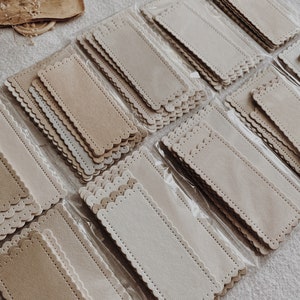 Puede incluir: Se ven varias pilas de etiquetas rectangulares de papel beige claro con bordes festoneados. Cada etiqueta tiene un borde cosido y est&aacute; empaquetada en pl&aacute;stico transparente. Las etiquetas est&aacute;n dispuestas sobre una superficie clara y texturizada.