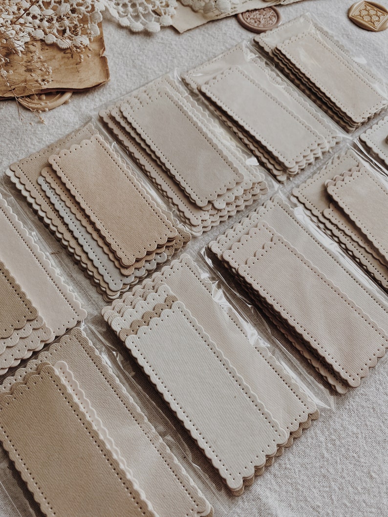 Puede incluir: Se ven varias pilas de etiquetas rectangulares de papel beige claro con bordes festoneados. Cada etiqueta tiene un borde cosido y est&aacute; empaquetada en pl&aacute;stico transparente. Las etiquetas est&aacute;n dispuestas sobre una superficie clara y texturizada.