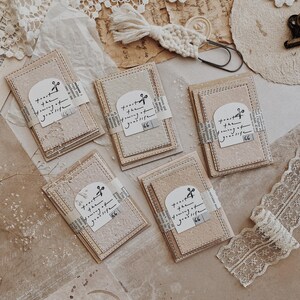 May include: Stacks of beige and pink paper cards with stitched edges are arranged on a textured surface. Each card is wrapped with a strip of paper featuring text and a musical note. A macrame accent and lace trim are also visible.