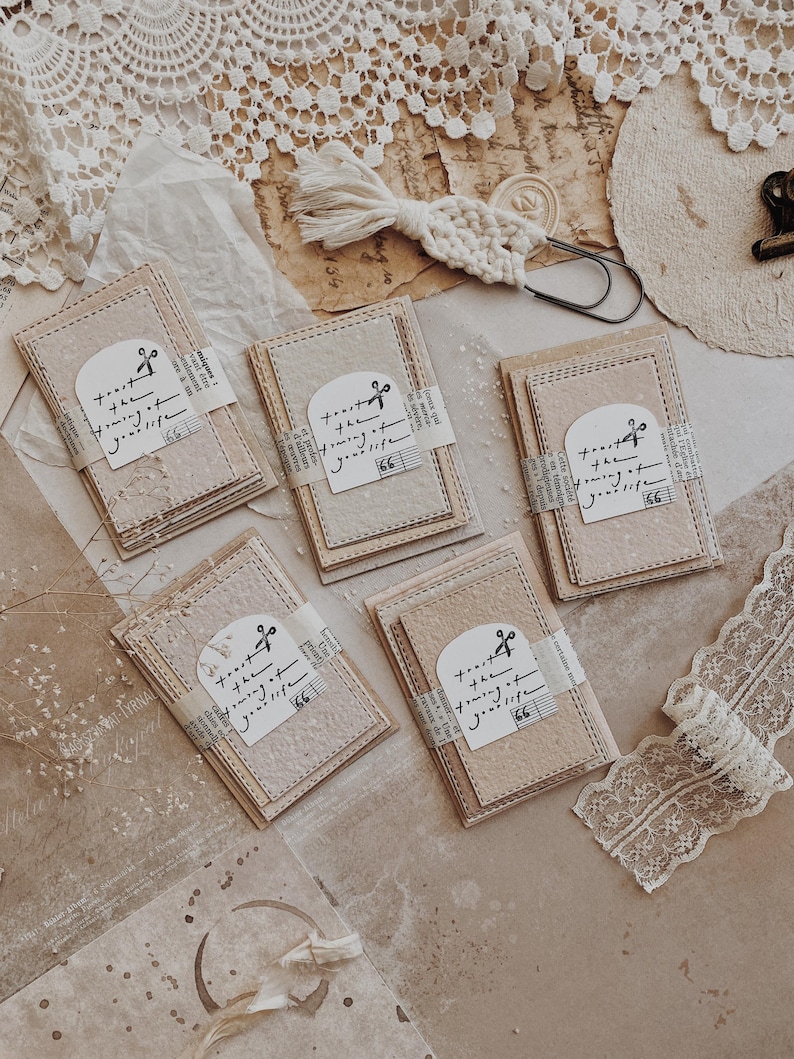 May include: Stacks of beige and pink paper cards with stitched edges are arranged on a textured surface. Each card is wrapped with a strip of paper featuring text and a musical note. A macrame accent and lace trim are also visible.