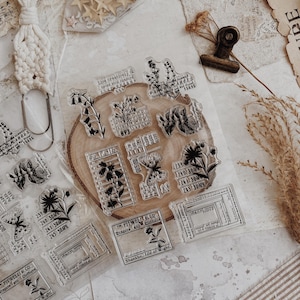 May include: A collection of clear stamps with botanical and label designs, including text like "FITWELL & CO." and "PARCEL LABELS." The stamps are arranged on a wooden slice and paper, with decorative elements like stars, lace, and a macrame clip.