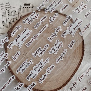 Könnte beinhalten: Ein transparenter Aufkleberbogen mit verschiedenen handgeschriebenen Phrasen in dunkelbrauner Tinte, darunter "create try new things", "you just need to breathe", "memory collecting", "lovely day", "happy moments", "good things are coming" und "inspired love".