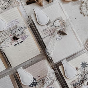 May include: A collection of rectangular journals featuring cream-colored paper with diagonal lines, secured by white teardrop-shaped magnetic clasps. Each journal is wrapped in white lace ribbon and adorned with metal charms and vintage-style stamps.